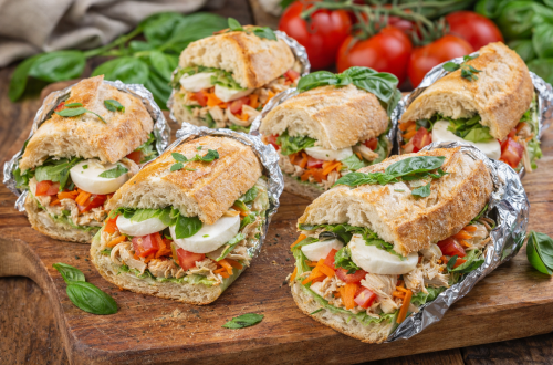 Gefülltes Baguette mit Hähnchenbrust, Mozzarella, Tomaten, Möhren, Eisbergsalat und frischem Basilikum auf einem rustikalen Holztisch, in Scheiben geschnitten, ohne Text, im Querformat.