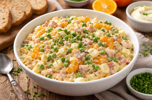 Cremiger Eiersalat mit Schinken, Erbsen und Mandarinen in einer weißen Schüssel auf Holztisch.