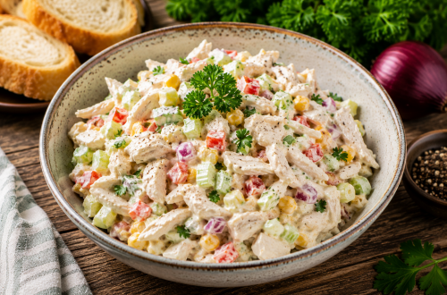 Cremiger Geflügelsalat mit zarten Hähnchenstreifen, Paprika, Sellerie, Mais und roten Zwiebeln in würzigem Dressing, serviert in einer rustikalen Schale mit frischen Kräutern.