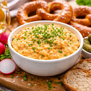 Cremiger bayerischer Obatzter in einer weißen Schüssel, mit Schnittlauch bestreut, serviert mit Brezeln, Brot, Radieschen, Gewürzgurken und einem Bier.