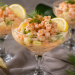 Krabben-Spargelcocktail in eleganten Cocktailgläsern mit zarten Garnelen, weißem Spargel, cremigem Dressing, frischem Dill und Zitronenscheiben. Arrangiert auf einem festlich gedeckten Tisch mit Spargelstangen und Kräutern. Fotorealistische Darstellung einer klassischen Vorspeise mit Krabben und Spargel.