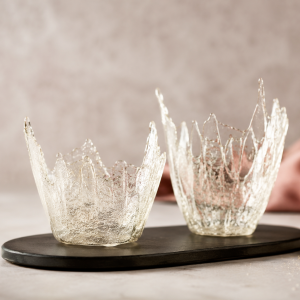 Fotorealistische Darstellung von essbarem Glas aus karamellisiertem Zucker in Form von transparenten, glasähnlichen Schalen auf einer dunklen Servierplatte. Die glänzende Zuckerdekoration wird in weichem Licht präsentiert und eignet sich als elegante Dekoration für Desserts und Torten.
