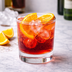 Fotorealistisches Bild eines Americano-Cocktails mit Eiswürfeln in einem Tumblerglas, dekoriert mit Orangenscheiben, auf heller Oberfläche mit unscharfem Barhintergrund.