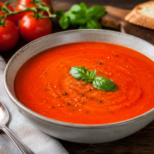 Cremige, leuchtend rote Tomatensuppe in einer hellen Keramikschale, garniert mit frischem Basilikum und schwarzem Pfeffer. Daneben liegen reife Tomaten, Basilikumblätter, rustikales Weißbrot und ein Löffel auf einem dunklen Holztisch. Fotorealistische Food-Fotografie in warmer, natürlicher Beleuchtung.