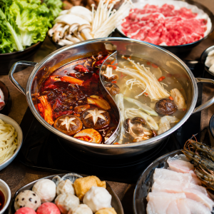 Großer chinesischer Hot Pot mit geteilter Brühe auf einem Tisch, umgeben von frischem Gemüse, Enoki-Pilzen, Tofu, dünn geschnittenem Rindfleisch, Garnelen, Weißfisch, Nudeln und verschiedenen Dips. Die eine Brühe ist scharf und rot, die andere mild und pilzbetont. Das Arrangement zeigt einen einladenden Hot-Pot-Abend für mehrere Personen mit vielfältigen Zutaten zum gemeinsamen Kochen.