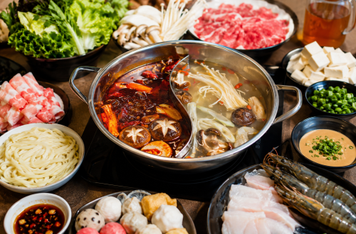 Großer chinesischer Hot Pot mit geteilter Brühe auf einem Tisch, umgeben von frischem Gemüse, Enoki-Pilzen, Tofu, dünn geschnittenem Rindfleisch, Garnelen, Weißfisch, Nudeln und verschiedenen Dips. Die eine Brühe ist scharf und rot, die andere mild und pilzbetont. Das Arrangement zeigt einen einladenden Hot-Pot-Abend für mehrere Personen mit vielfältigen Zutaten zum gemeinsamen Kochen.