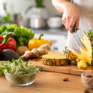 Frische Ananas wird auf einem Holzbrett in einer hellen Küche geschnitten, umgeben von Papaya, Kiwi, Avocado, Paprika, Ingwer und weiteren frischen Zutaten. Das fotorealistische Bild veranschaulicht die Wirkung natürlicher Enzyme in Lebensmitteln und deren Einsatz in der Küche.