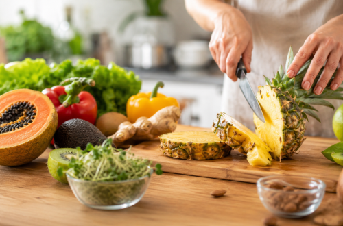Frische Ananas wird auf einem Holzbrett in einer hellen Küche geschnitten, umgeben von Papaya, Kiwi, Avocado, Paprika, Ingwer und weiteren frischen Zutaten. Das fotorealistische Bild veranschaulicht die Wirkung natürlicher Enzyme in Lebensmitteln und deren Einsatz in der Küche.