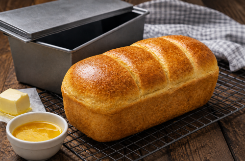 Frisch gebackenes Dinkel-Toastbrot mit goldbrauner, glänzender Kruste auf einem schwarzen Kuchengitter. Daneben stehen eine Toastbrotform, geschmolzene Butter und ein Stück Butter auf einem rustikalen dunklen Holztisch. Perfektes selbst gebackenes Dinkel-Toastbrot für Frühstück und Sandwiches.
