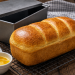 Frisch gebackenes Dinkel-Toastbrot mit goldbrauner, glänzender Kruste auf einem schwarzen Kuchengitter. Daneben stehen eine Toastbrotform, geschmolzene Butter und ein Stück Butter auf einem rustikalen dunklen Holztisch. Perfektes selbst gebackenes Dinkel-Toastbrot für Frühstück und Sandwiches.