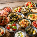 Frisch angerichtetes internationales Tapas-Buffet auf einem rustikalen Holztisch mit verschiedenen Häppchen, darunter Oliven, Hummus, Garnelen, Fleischbällchen in Tomatensauce, gefüllte Weinblätter, Antipasti, Käse, luftgetrockneter Schinken und frisches Baguette. Farbenfrohe Auswahl an Vorspeisen aus verschiedenen Ländern für ein geselliges Buffet.