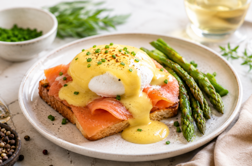 Egg Benedict mit Räucherlachs und grünem Spargel auf knusprigem Toast, serviert auf einem hellen Keramikteller. Das pochierte Ei ist mit cremiger Sauce Hollandaise überzogen und mit frischem Schnittlauch garniert. Daneben liegen gebratene Spargelstangen, Pfefferkörner und frische Kräuter. Helles, natürliches Food-Foto in hochwertiger Brunch-Optik.