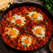 Shakshuka in einer gusseisernen Pfanne mit Eiern, würziger Tomatensauce, frischer Petersilie und Fladenbrot auf rustikalem Holztisch.