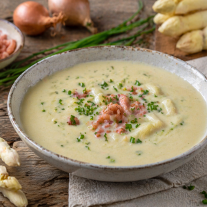 Cremige Spargelcremesuppe mit weißen Spargelstücken, Krabben und frischem Schnittlauch in einer Schale, angerichtet auf rustikalem Holztisch mit frischem Spargel und Zutaten im Hintergrund