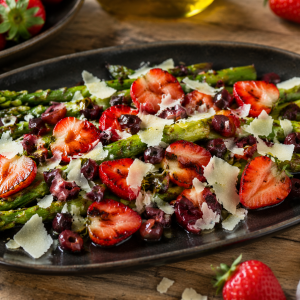 Gegrillter grüner Spargel auf einer dunklen Platte, serviert mit halbierten, leicht angerösteten Erdbeeren, gehackten schwarzen Oliven und frisch gehobeltem Parmesan, angerichtet auf einem rustikalen Holztisch.