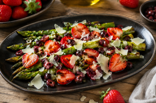 Gegrillter grüner Spargel auf einer dunklen Platte, serviert mit halbierten, leicht angerösteten Erdbeeren, gehackten schwarzen Oliven und frisch gehobeltem Parmesan, angerichtet auf einem rustikalen Holztisch.