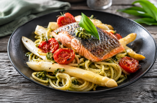 Fotorealistisches Gericht mit knusprig gebratener Lachsforelle auf Bärlauchspaghetti, dazu weißer Spargel und halbierte, leicht geröstete Cherrytomaten auf einem rustikalen Teller.