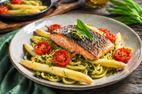 Fotorealistisches Gericht mit knusprig gebratener Lachsforelle auf Bärlauchspaghetti, dazu weißer Spargel und halbierte, leicht geröstete Cherrytomaten auf einem rustikalen Teller.