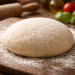 Frischer ungebackener Pizzateig als Teigkugel auf bemehltem Holzbrett in rustikaler Küche mit Nudelholz und Zutaten im Hintergrund – Vorbereitung für hausgemachte Pizza nach direkter Teigführung.