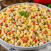 Cremiger Hörnchen-Nudelsalat mit Lyoner Fleischkäse, Erbsen, Mais, Paprika, Cornichons und Mini-Tomaten in einer großen weißen Schüssel auf rustikalem Holztisch dekoriert.