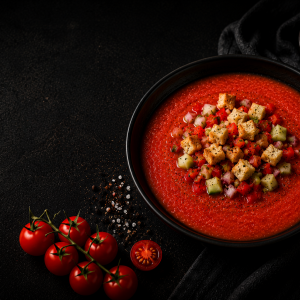 Fotorealistisches Beitragsbild einer klassischen Gazpacho in einer schwarzen Schüssel auf dunklem Hintergrund. Die kalte spanische Tomatensuppe ist mit knusprigen Croutons sowie fein gewürfelten Tomaten, Gurken und roten Zwiebeln garniert. Daneben liegen frische Cherrytomaten, grobes Meersalz und eine kleine Schale mit Croutons in einer edlen, stimmungsvoll beleuchteten Food-Fotografie.