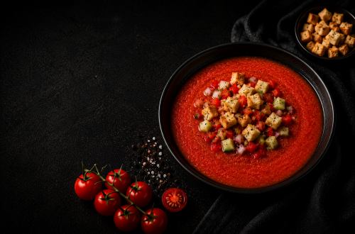 Fotorealistisches Beitragsbild einer klassischen Gazpacho in einer schwarzen Schüssel auf dunklem Hintergrund. Die kalte spanische Tomatensuppe ist mit knusprigen Croutons sowie fein gewürfelten Tomaten, Gurken und roten Zwiebeln garniert. Daneben liegen frische Cherrytomaten, grobes Meersalz und eine kleine Schale mit Croutons in einer edlen, stimmungsvoll beleuchteten Food-Fotografie.