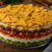 Schichtsalat Chili con Carne mit Hackfleisch, Kidneybohnen, Mais, Salsa, Eisbergsalat, Creme fraîche, geriebenem Gouda und Nachos in einer Glasschüssel auf rustikalem Holztisch