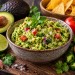 Frisch zubereitete Guacamole mit Avocado, Tomaten und roten Zwiebeln in einer rustikalen Schale, serviert mit Tortilla-Chips, Limette und Avocado.