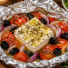 Grillfeta in Alufolie mit Tomaten, roten Zwiebeln, schwarzen Oliven, Olivenöl und Oregano, frisch vom Grill serviert.